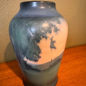 Peut inclure: Un vase en céramique bleu et vert avec un corps large et un col étroit. Le vase a une surface texturée avec un motif de paysage dans les tons de bleu, vert et blanc. Le vase est posé sur une surface en bois.