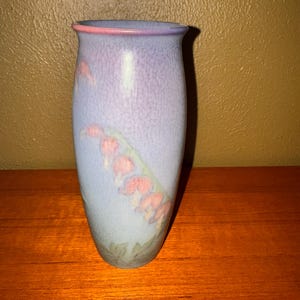 May include: A ceramic vase with a light blue and purple gradient. The vase features a floral design with pink and green accents. The rim is trimmed with pink. The vase is displayed on a wooden surface.