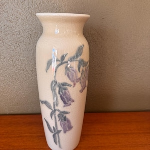 May include: A cream-colored ceramic vase with a floral design. The vase features a slender, elongated shape with a slightly flared rim. The floral pattern includes purple bell-shaped flowers and green leaves. The vase is approximately 8 inches tall.