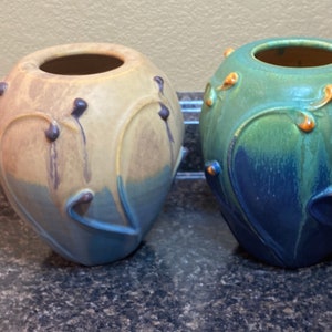 May include: Two ceramic vases with a heart-shaped design. The vase on the left is yellow, blue, and brown. The vase on the right is green, blue, and brown.