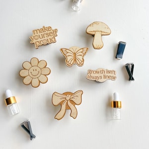 Könnte beinhalten: Sortiment an Holz-Lufterfrischern für Autos mit verschiedenen Designs, darunter ein Schmetterling, ein Pilz, eine Blume, eine Schleife und ein Textdesign mit der Aufschrift "make yourself proud" und "growth isn't always linear".