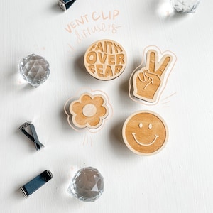 Könnte beinhalten: Ein Set aus fünf Holz-Lufterfrischern für das Auto mit verschiedenen Designs. Die Designs umfassen eine Blume, ein Smiley, ein Peace-Zeichen und einen Kreis mit dem Text "Faith Over Fear". Die Lufterfrischer sind an schwarzen Lüftungsclips für das Auto befestigt.