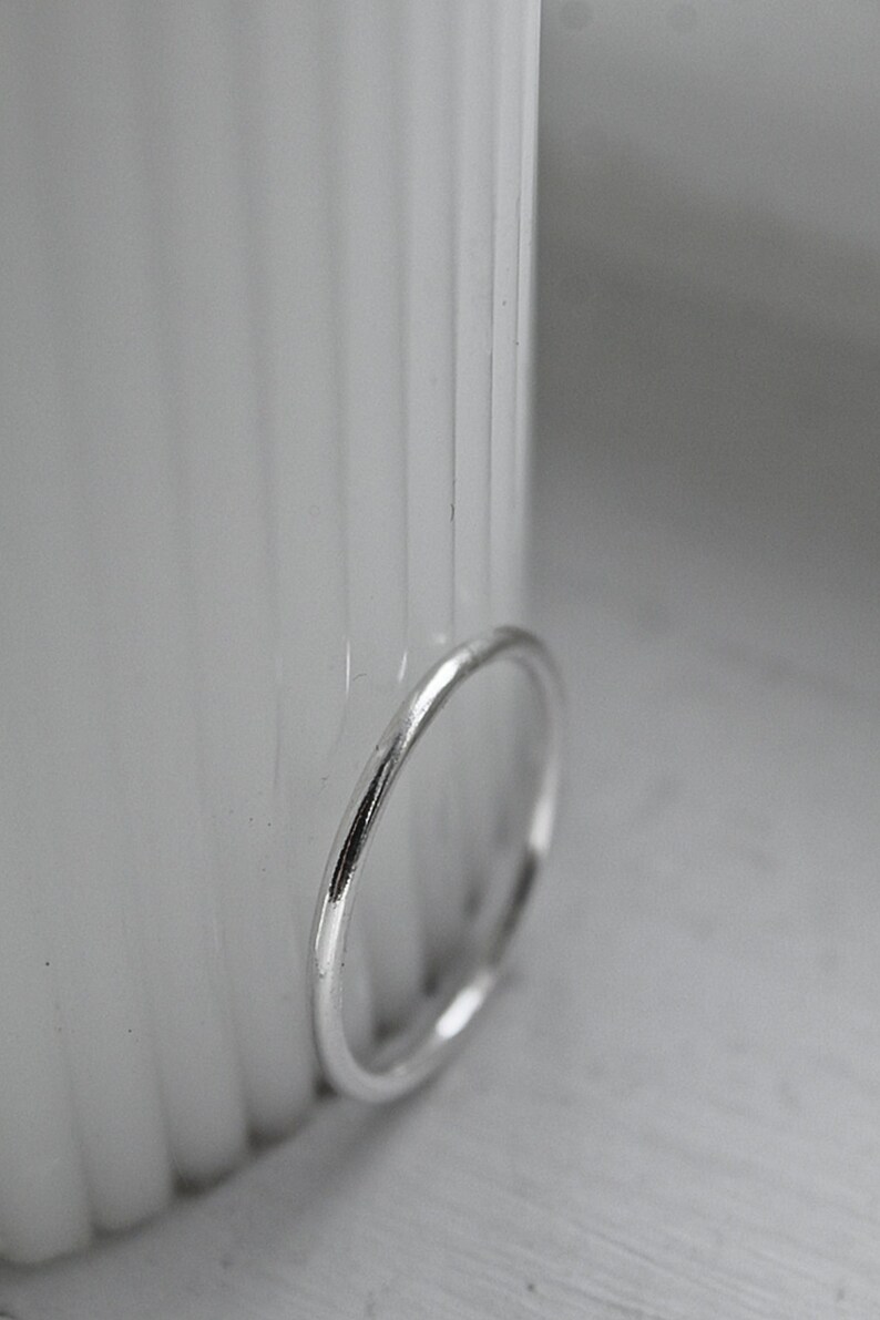 May include: A simple silver ring with a polished surface. The ring is positioned against a blurred white backdrop, accentuating its understated, elegant design. This piece of jewellery is suitable for everyday wear.