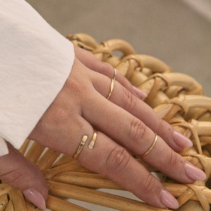 K&ouml;nnte beinhalten: Eine Hand tr&auml;gt vier goldene Ringe, darunter einen einzigartigen Doppelring, auf einer geflochtenen Weidenoberfl&auml;che.