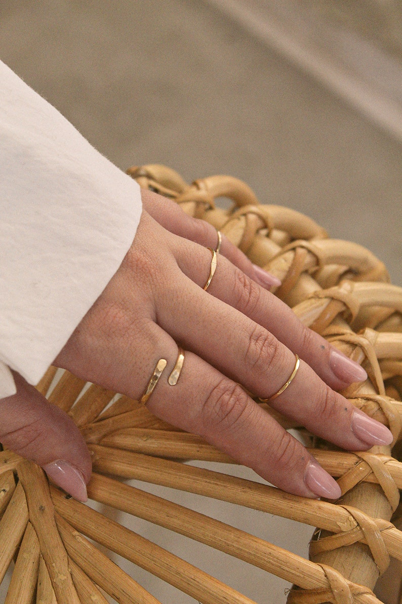 K&ouml;nnte beinhalten: Eine Hand tr&auml;gt vier goldene Ringe, darunter einen einzigartigen Doppelring, auf einer geflochtenen Weidenoberfl&auml;che.
