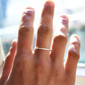 May include: A slender, silver ring worn on a finger. The ring has a minimalist design, with a smooth, polished surface. The hand is in focus, with the ring on the ring finger. The background is blurred, with natural light.
