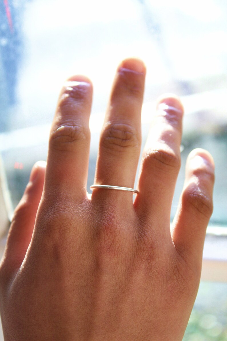 May include: A slender, silver ring worn on a finger. The ring has a minimalist design, with a smooth, polished surface. The hand is in focus, with the ring on the ring finger. The background is blurred, with natural light.