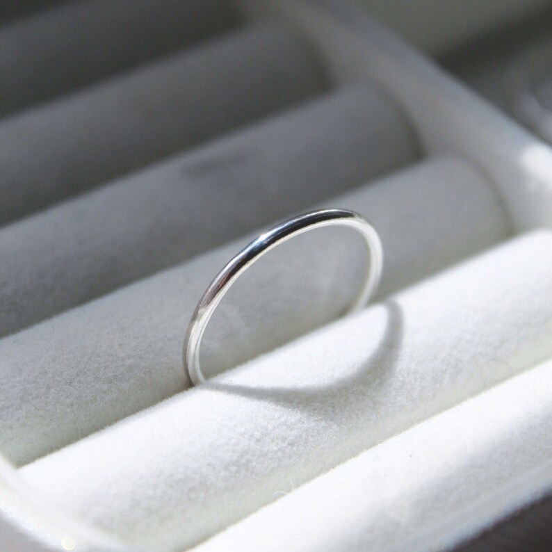 May include: A slender, silver ring with a shiny surface. The ring is displayed against a white, textured backdrop, likely within a jewellery box. The ring's minimalist design suggests a simple, elegant style, suitable for daily wear.