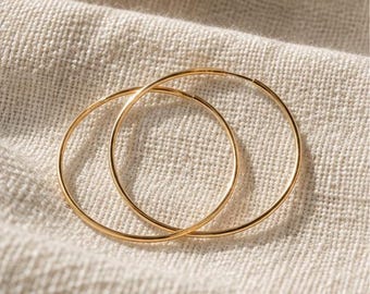 Op de afbeelding: Een paar dunne, ronde gouden oorringen met een gepolijste afwerking. De oorbellen zijn eenvoudig, lichtgewicht en hebben een doorlopend draadontwerp dat elkaar licht overlapt, wat zorgt voor een klassieke en ingetogen uitstraling voor elke collectie.