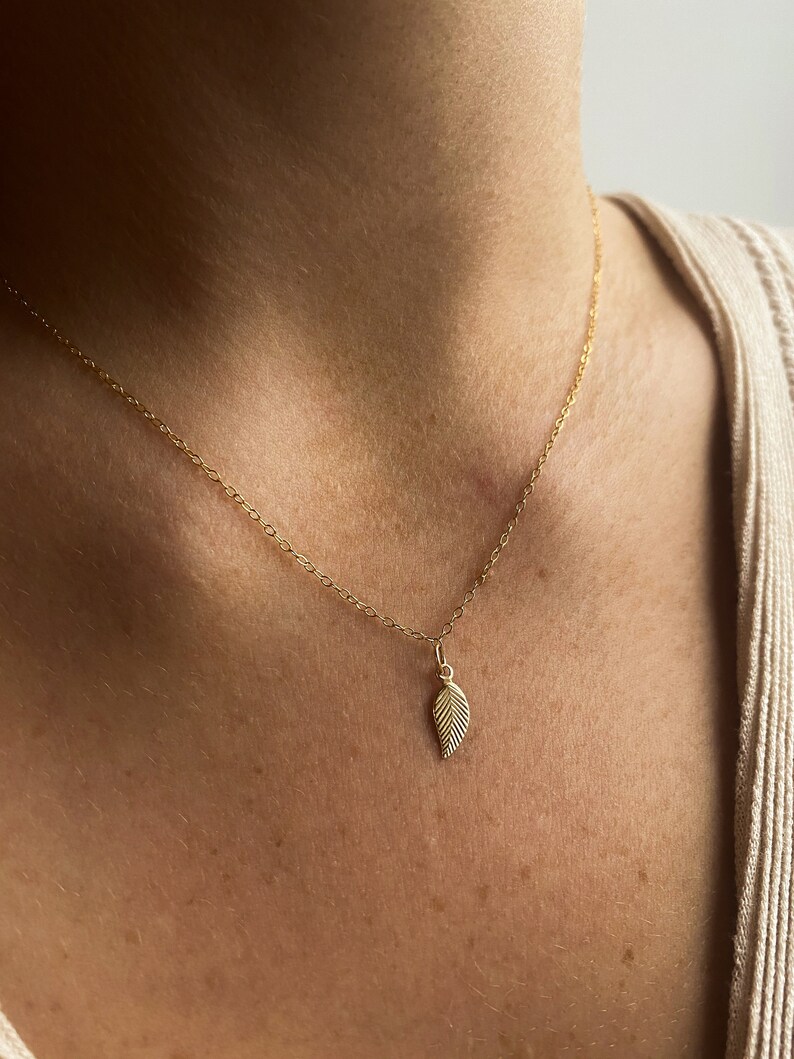 May include: A delicate gold chain necklace with a small leaf pendant. The leaf is detailed with fine lines and has a slightly textured surface.