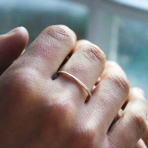 May include: A slender, silver ring adorns a finger. The ring is a plain band, reflecting light. The hand's skin tone is medium, with visible texture. The background is blurred, suggesting a window.