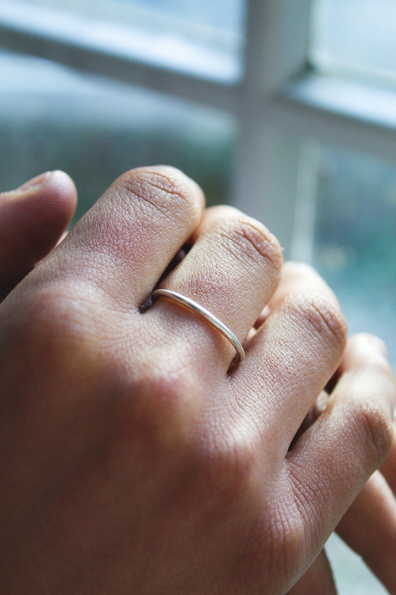 May include: A slender, silver ring adorns a finger. The ring is a plain band, reflecting light. The hand's skin tone is medium, with visible texture. The background is blurred, suggesting a window.