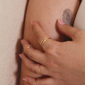 K&ouml;nnte beinhalten: Eine Hand tr&auml;gt vier goldene Ringe, einer davon ist ein gebogenes Band. Die Hand ruht auf dem Arm einer Person.