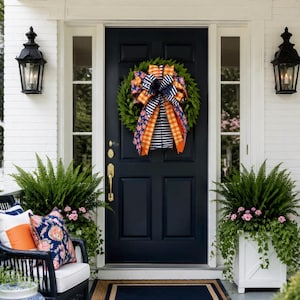 May include: A green wreath hangs on a black front door, featuring a large decorative bow with orange plaid, navy blue and white striped, and navy blue floral patterned ribbons. The door is flanked by white planters with ferns and flowers.
