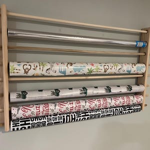 May include: A wooden wrapping paper organizer with multiple rolls of gift wrap. The rolls feature various patterns, including jungle animals, trucks, and holiday-themed text. The organizer is mounted on a light gray wall.