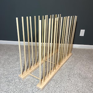 May include: A wooden plate rack with multiple vertical dowels. The rack is made of light-colored wood and is designed to hold plates upright. The rack is on a carpeted floor with a dark gray wall in the background.