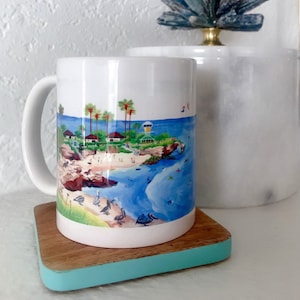 May include: White ceramic mug with a colorful illustration of a beach scene with palm trees, a lifeguard tower, and birds. The mug is on a wooden coaster with a turquoise border.