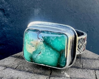 Puede incluir: Un anillo de plata con una piedra grande, rectangular, de color verde esmeralda. El anillo tiene una banda gruesa con grabados decorativos. La piedra está engastada en un bisel de plata.