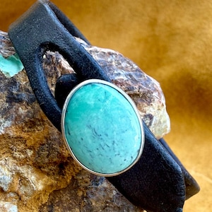 May include: Black leather bracelet featuring a large, oval turquoise stone with a silver bezel. The bracelet has a unique, open design. The turquoise stone has a mottled pattern and is a vibrant shade of blue-green.