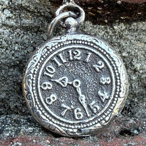 Puede incluir: Colgante de reloj de bolsillo plateado con un diseño ornamentado. La esfera del reloj muestra los números del 1 al 12, con las manecillas que indican aproximadamente las 9:26. El reloj tiene un lazo en la parte superior para sujetarlo a una cadena.
