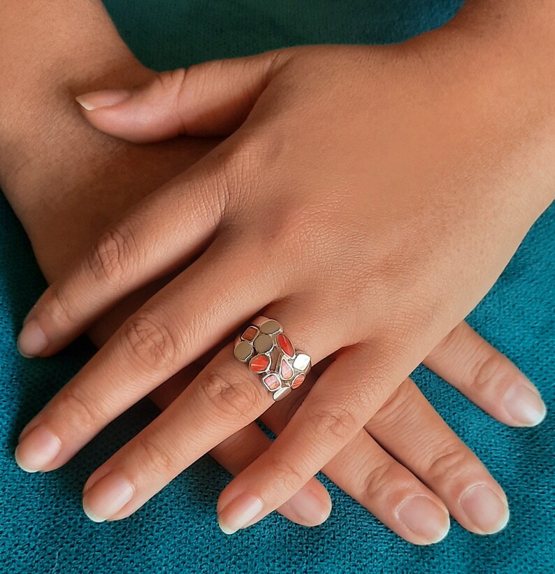 May include: A silver ring with a multi-colored design. The ring is made up of small, oval-shaped pieces of stone in shades of pink, orange, and white. The ring is worn on a person's finger.