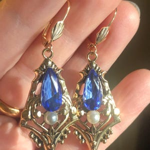 Op de afbeelding: Een paar oorbellen in vintage-stijl met een goudkleurig filigraan ontwerp. Elke oorbel heeft een grote, druppelvormige blauwe edelsteen en een kleine, ronde parel. Met oorhaak sluiting.
