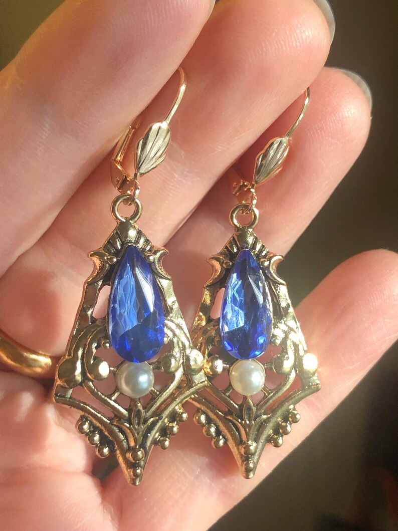 Op de afbeelding: Een paar oorbellen in vintage-stijl met een goudkleurig filigraan ontwerp. Elke oorbel heeft een grote, druppelvormige blauwe edelsteen en een kleine, ronde parel. Met oorhaak sluiting.