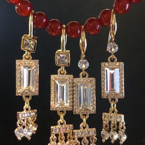 May include: A necklace with red beads and four pairs of gold earrings. Each earring has a rectangular, clear stone surrounded by smaller clear stones. Some earrings have dangling clear stones.