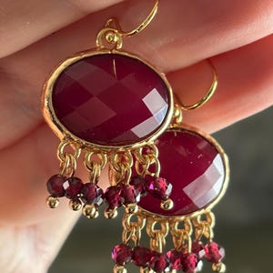 May include: Gold-coloured earrings featuring faceted, oval burgundy stones. Small burgundy beads hang from the bottom of each earring. The earrings are suspended by gold-coloured hooks.