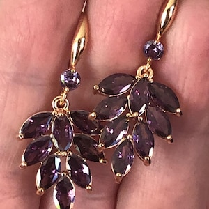 Puede incluir: Pendientes colgantes dorados con racimos de piedras preciosas moradas profundas talladas en marquesa. Cada pendiente tiene una pequeña piedra preciosa morada redonda en la parte superior. El diseño se asemeja a un arreglo floral estilizado.