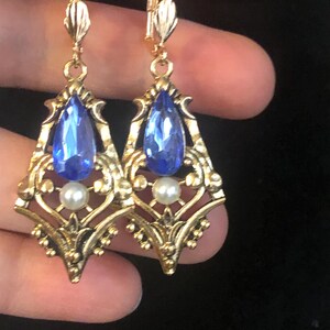 Op de afbeelding: Een paar goudkleurige oorbellen met een blauwe, druppelvormige edelsteen en een kleine witte parel. De oorbellen hebben een sierlijk filigraanontwerp en zijn bevestigd aan een klapbeugel. Een rode kralenketting is zichtbaar op de achtergrond.