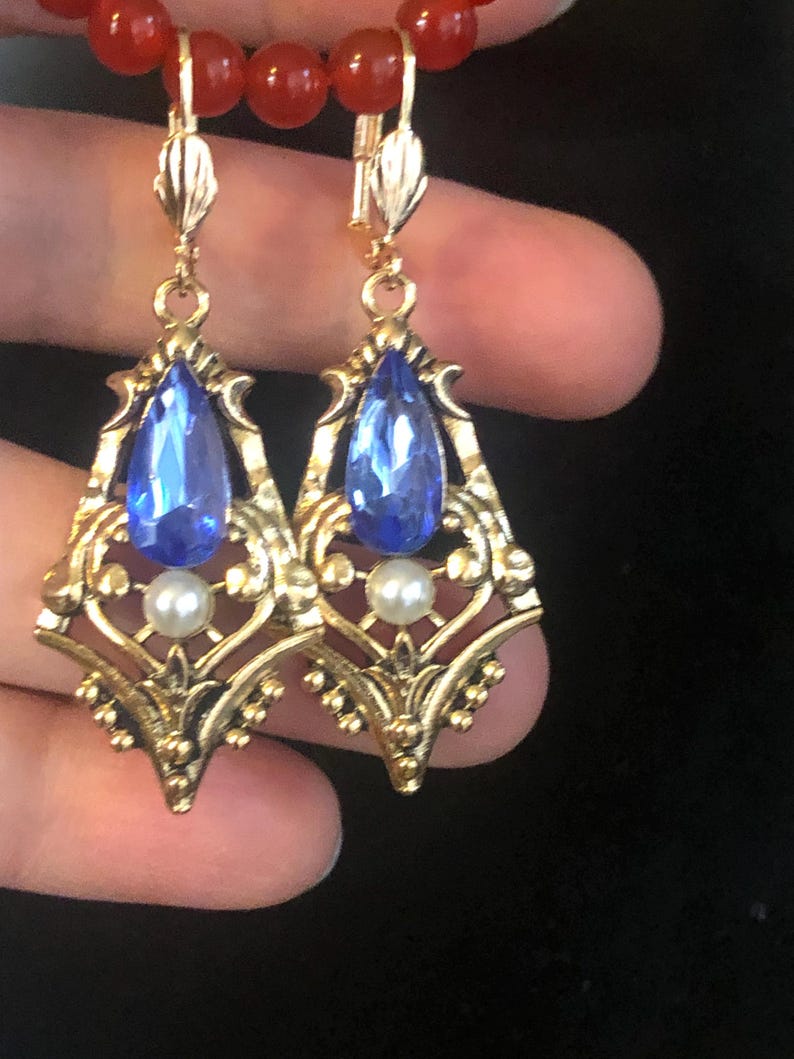 Op de afbeelding: Een paar goudkleurige oorbellen met een blauwe, druppelvormige edelsteen en een kleine witte parel. De oorbellen hebben een sierlijk filigraanontwerp en zijn bevestigd aan een klapbeugel. Een rode kralenketting is zichtbaar op de achtergrond.