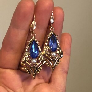Op de afbeelding: Een paar goudkleurige oorbellen met een druppelvormige, diepblauwe edelsteen en een kleine parel. De oorbellen hebben een sierlijk filigraanontwerp en een decoratieve haaksluiting. De oorbellen worden in een hand gehouden.