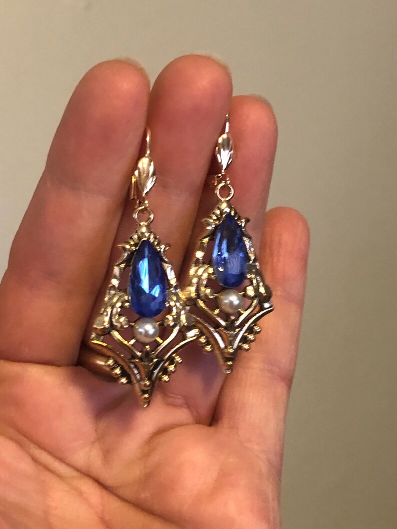 Op de afbeelding: Een paar goudkleurige oorbellen met een druppelvormige, diepblauwe edelsteen en een kleine parel. De oorbellen hebben een sierlijk filigraanontwerp en een decoratieve haaksluiting. De oorbellen worden in een hand gehouden.