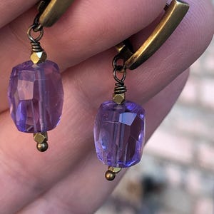 May include: A pair of earrings with a brass hoop and a dangling purple gemstone. The gemstone is faceted and has a light purple hue.