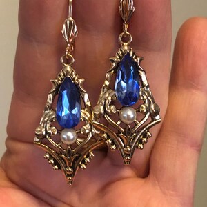 Op de afbeelding: Een paar goudkleurige oorbellen met een vintage design. Elke oorbel heeft een grote, druppelvormige blauwe edelsteen, een kleine witte parel en sierlijke filigraan details. De oorbellen hebben een haaksluiting.