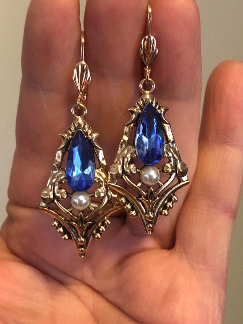 Op de afbeelding: Een paar goudkleurige oorbellen met een vintage design. Elke oorbel heeft een grote, druppelvormige blauwe edelsteen, een kleine witte parel en sierlijke filigraan details. De oorbellen hebben een haaksluiting.