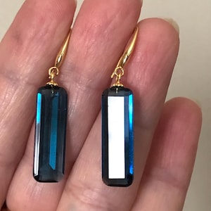 May include: A pair of gold-colored earrings with rectangular, dark teal-colored crystal pendants. The crystals have a vertical, light blue reflective stripe. The earrings are displayed against a neutral background.
