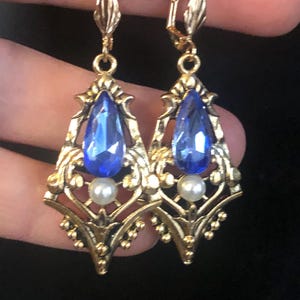 Op de afbeelding: Een paar goudkleurige oorhangers met een blauwe, druppelvormige edelsteen en een kleine witte parel. De oorbellen hebben een sierlijk filigraanontwerp en zijn bevestigd aan een klapbeugel. Rode kralen zijn zichtbaar op de achtergrond.