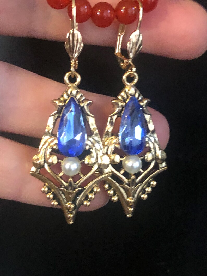 Op de afbeelding: Een paar goudkleurige oorhangers met een blauwe, druppelvormige edelsteen en een kleine witte parel. De oorbellen hebben een sierlijk filigraanontwerp en zijn bevestigd aan een klapbeugel. Rode kralen zijn zichtbaar op de achtergrond.
