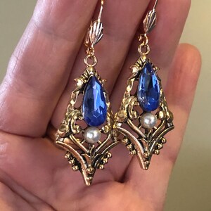 Op de afbeelding: Een paar oorbellen in vintage-stijl met een goudkleurig filigraanontwerp. Elke oorbel heeft een grote, druppelvormige blauwe edelsteen en een kleine, ronde witte parel. De oorbellen hebben een klapsluiting.