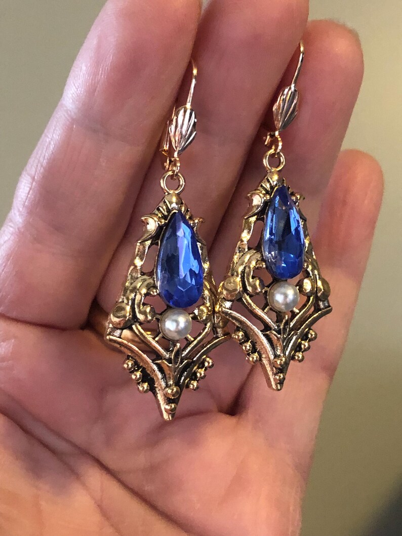 Op de afbeelding: Een paar oorbellen in vintage-stijl met een goudkleurig filigraanontwerp. Elke oorbel heeft een grote, druppelvormige blauwe edelsteen en een kleine, ronde witte parel. De oorbellen hebben een klapsluiting.