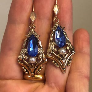 Op de afbeelding: Een paar goudkleurige oorhangers met een vintage uitstraling. Elke oorbel heeft een grote, druppelvormige blauwe edelsteen, een kleine witte parel en sierlijke filigraan details. De oorbellen hebben een haaksluiting.