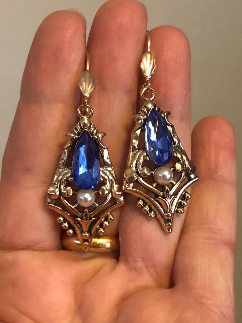 Op de afbeelding: Een paar goudkleurige oorhangers met een vintage uitstraling. Elke oorbel heeft een grote, druppelvormige blauwe edelsteen, een kleine witte parel en sierlijke filigraan details. De oorbellen hebben een haaksluiting.