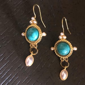 May include: A pair of gold-toned earrings with turquoise centers and pearl accents. Each earring features a teardrop-shaped pearl pendant. The earrings have a decorative design with a hook closure.