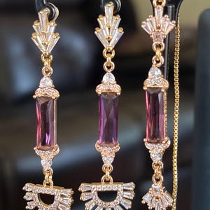 May include: A pair of gold-toned drop earrings featuring a vertical rectangular purple gemstone in the centre, accented with clear baguette and round crystals arranged in fan-shaped patterns at the top and bottom of each earring piece.