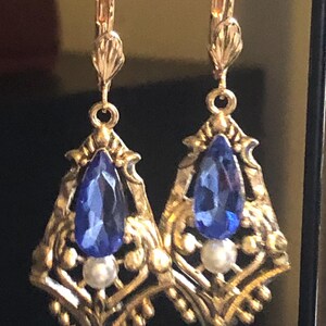 Op de afbeelding: Een paar goudkleurige oorbellen met een druppelvormige blauwe edelsteen en een kleine witte parel. De oorbellen hebben een sierlijk filigraanontwerp en een oorhaaksluiting. De oorbellen worden tegen een donkere achtergrond getoond.