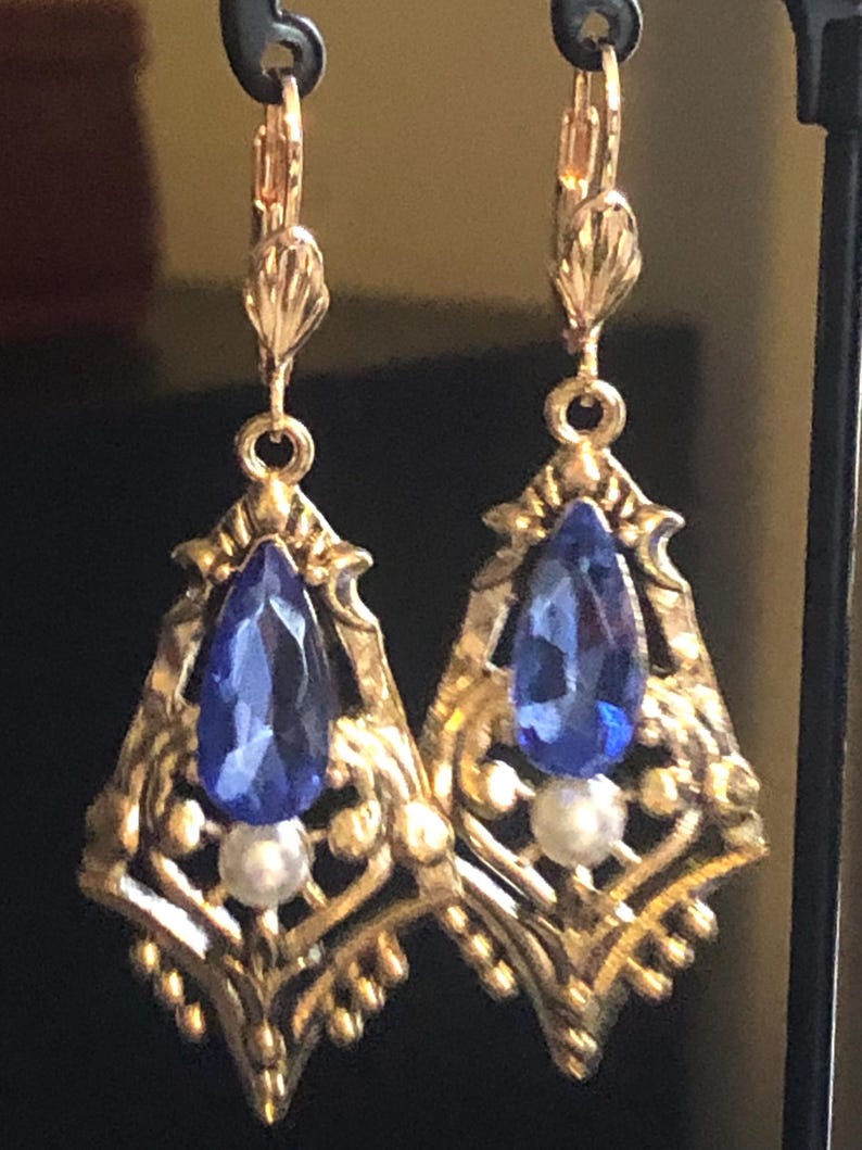 Op de afbeelding: Een paar goudkleurige oorbellen met een druppelvormige blauwe edelsteen en een kleine witte parel. De oorbellen hebben een sierlijk filigraanontwerp en een oorhaaksluiting. De oorbellen worden tegen een donkere achtergrond getoond.