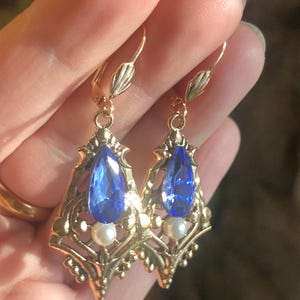 Op de afbeelding: Een paar goudkleurige oorhangers met sierlijke filigraan details. Elke oorbel heeft een grote, druppelvormige blauwe edelsteen en een kleine, ronde witte parel. De oorbellen hebben een klapsluiting.