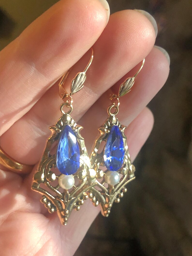 Op de afbeelding: Een paar goudkleurige oorhangers met sierlijke filigraan details. Elke oorbel heeft een grote, druppelvormige blauwe edelsteen en een kleine, ronde witte parel. De oorbellen hebben een klapsluiting.
